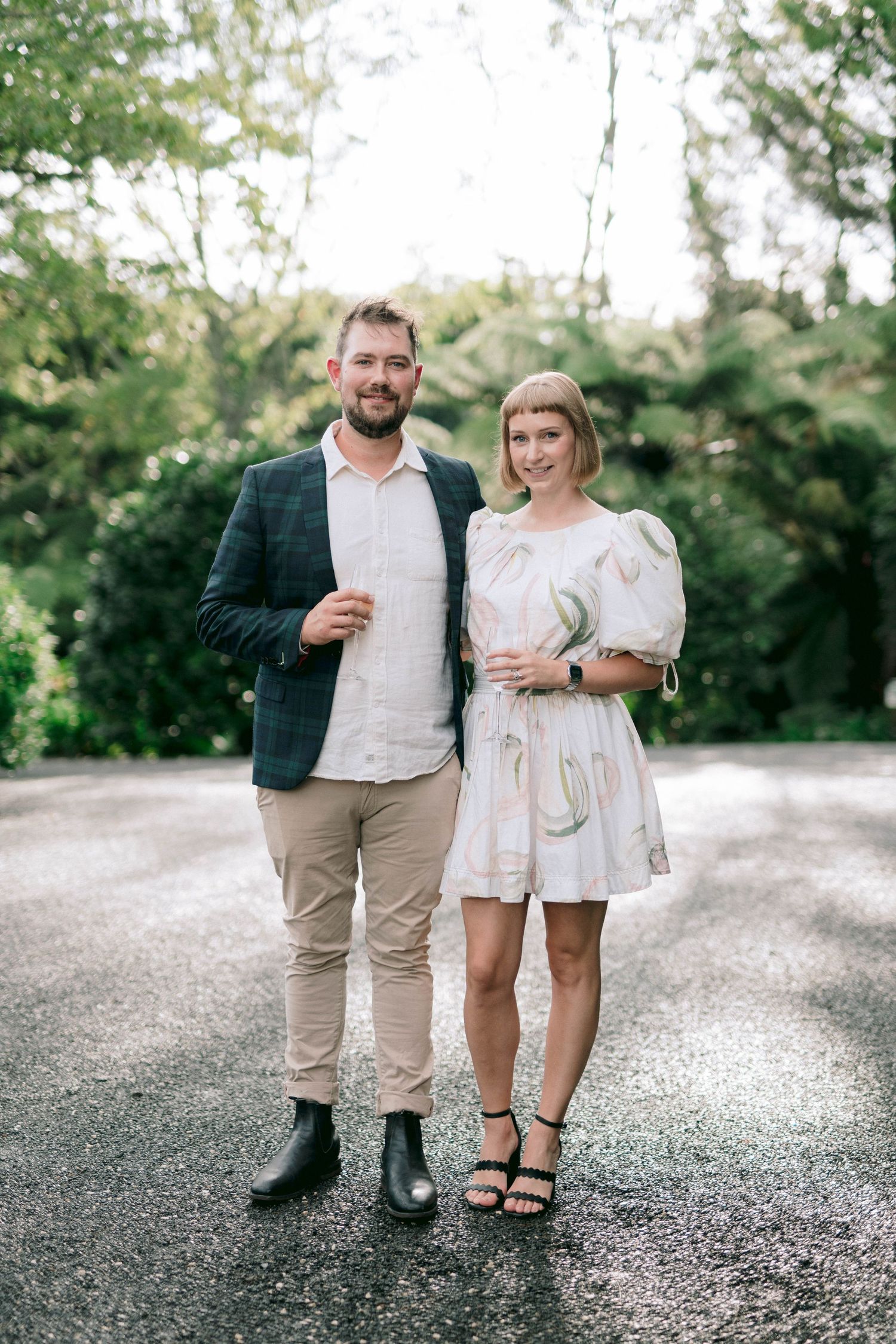 A couple in casual attire stands together on a gravel path surrounded by green foliage.