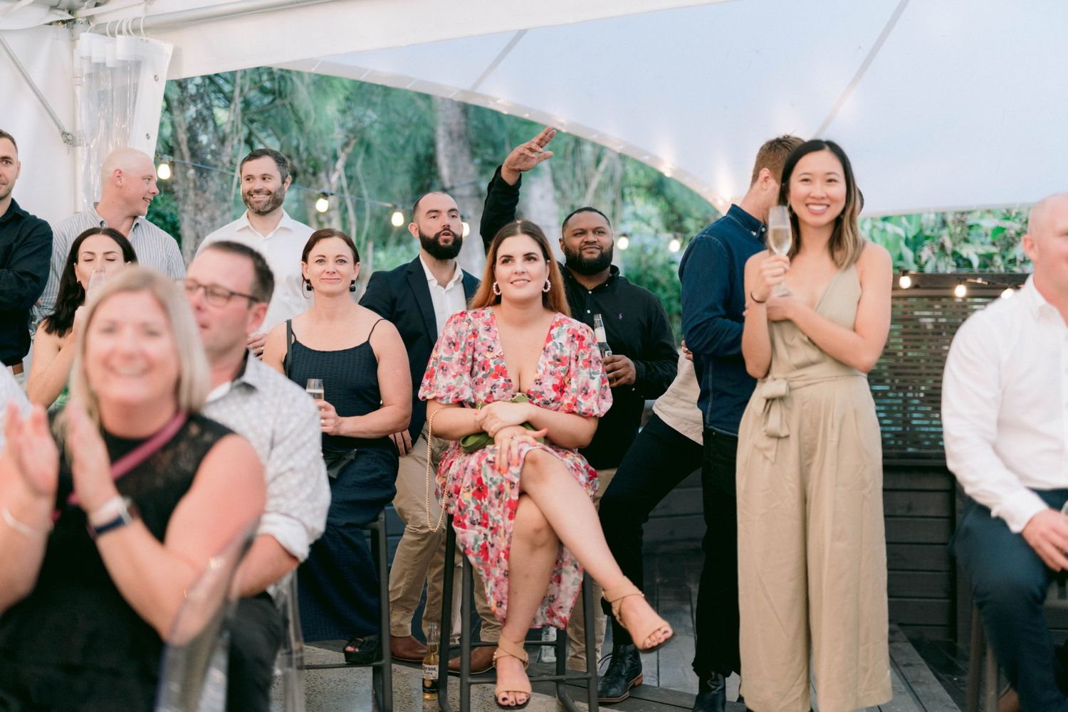 A group of wedding guests enjoying an outdoor reception under a white tent on a sunny day.