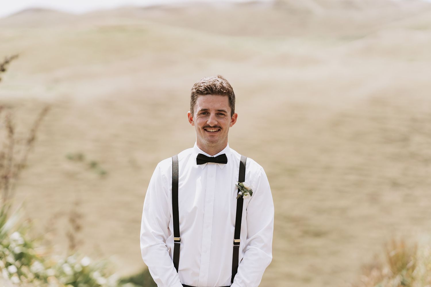 Someone in formal attire with a white shirt, black bow tie and black suspenders smiles in an outdoor setting.