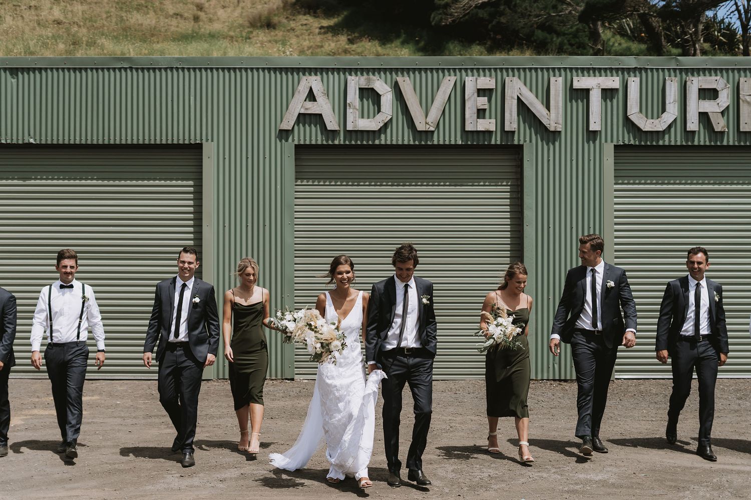 Wedding party poses in front of green warehouse doors with ADVENTURE sign during outdoor celebration photo shoot.