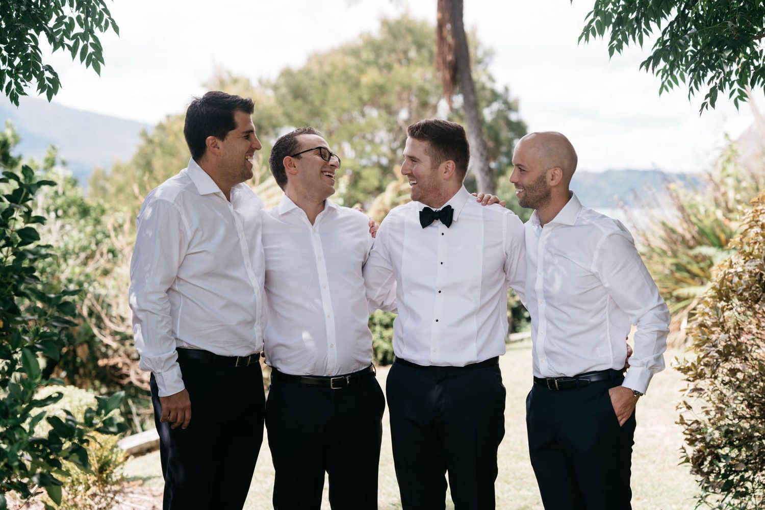 Group of groomsmen wearing white shirts and black pants share laughs at outdoor wedding venue with mountain backdrop.