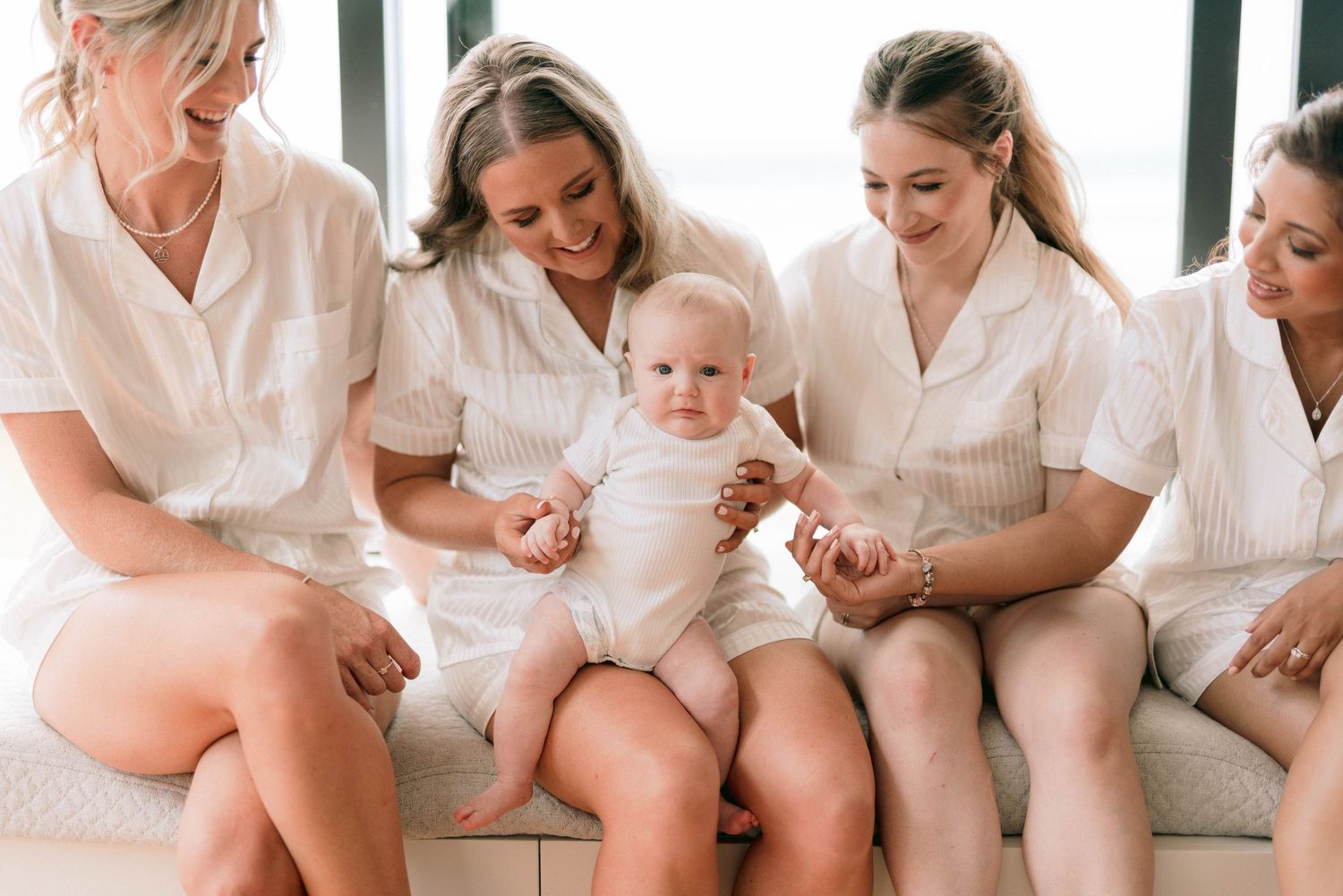 A baby wearing white sits surrounded by people in matching cream-colored outfits while gathering for a photo.