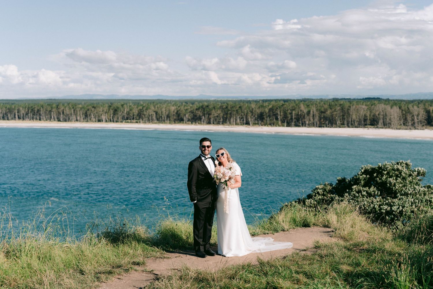 Wedding couple poses on coastal cliff overlooking pristine blue water and forested shoreline.