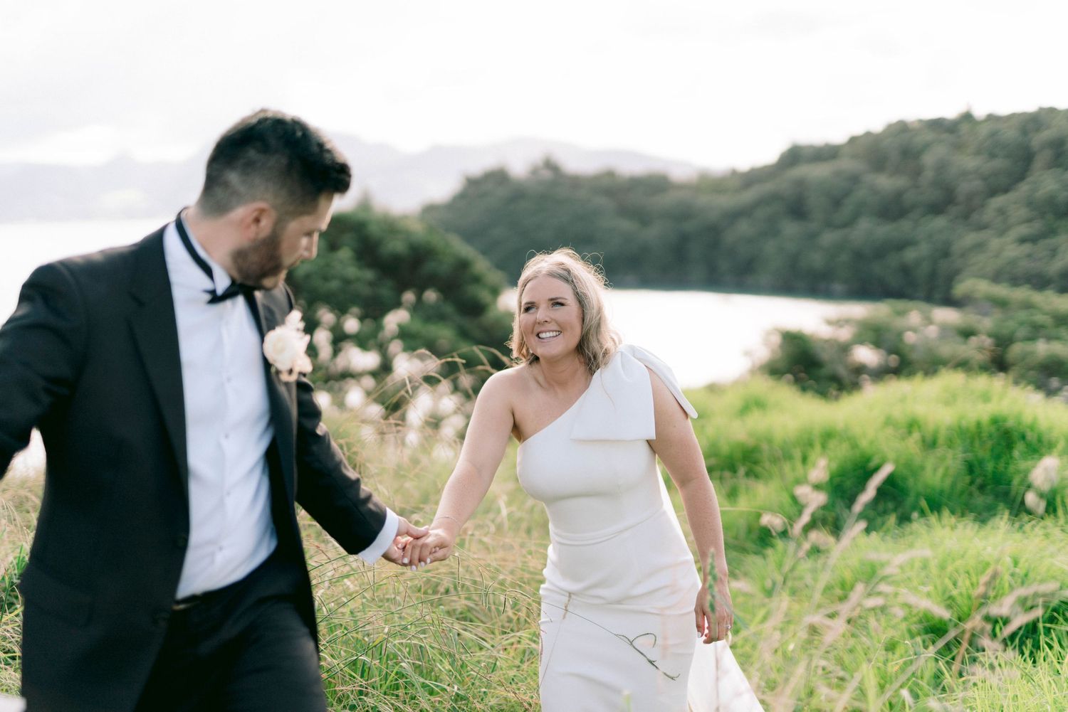 Happy couple walks through grassy hillside by the ocean in formal wedding attire.