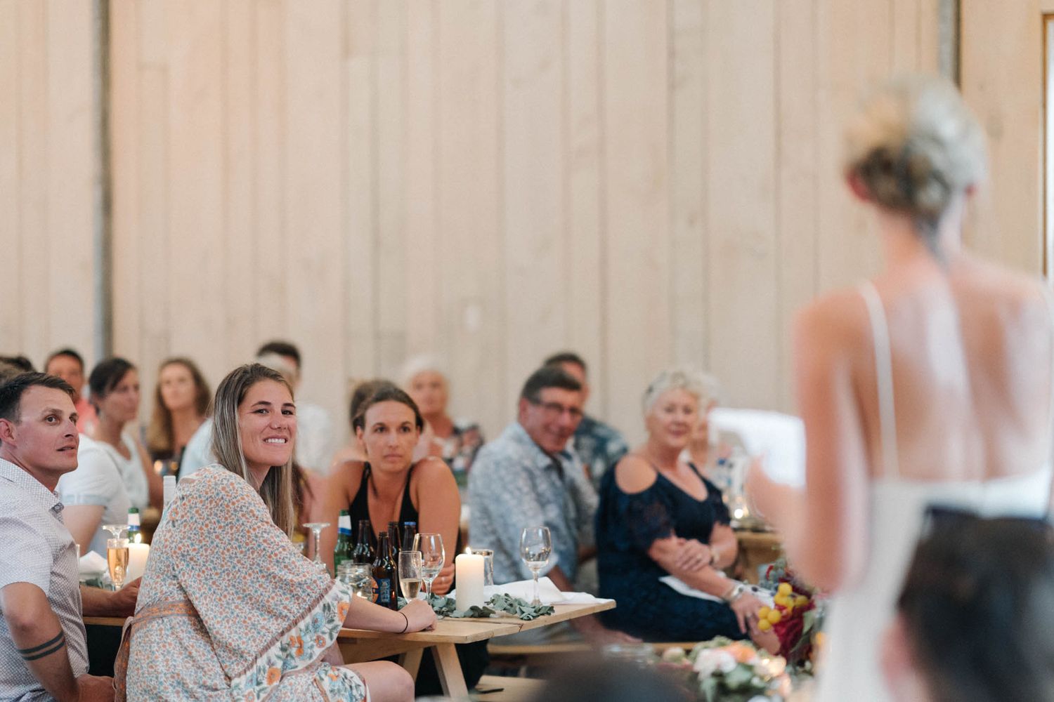 Wedding guests seated at tables listen attentively during a reception speech in a modern venue.