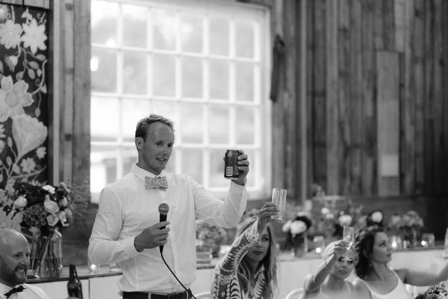 A wedding reception candid shot shows guests enjoying conversations and dining at decorated tables in a rustic venue.