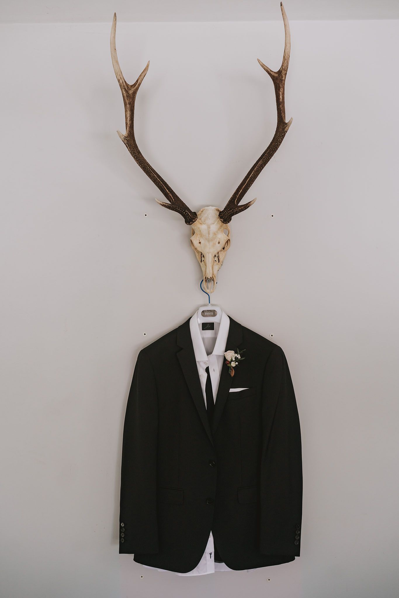 A black formal suit jacket and white dress shirt hanging below mounted deer antlers and skull on a white wall.