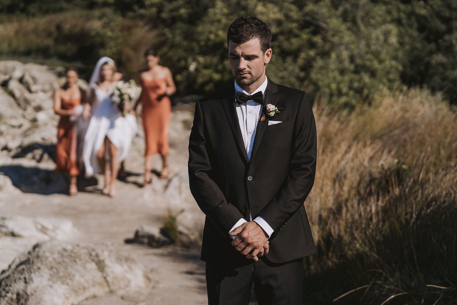 Wedding party stands on rock outcrop as groom waits pensively in black tuxedo during outdoor ceremony.
