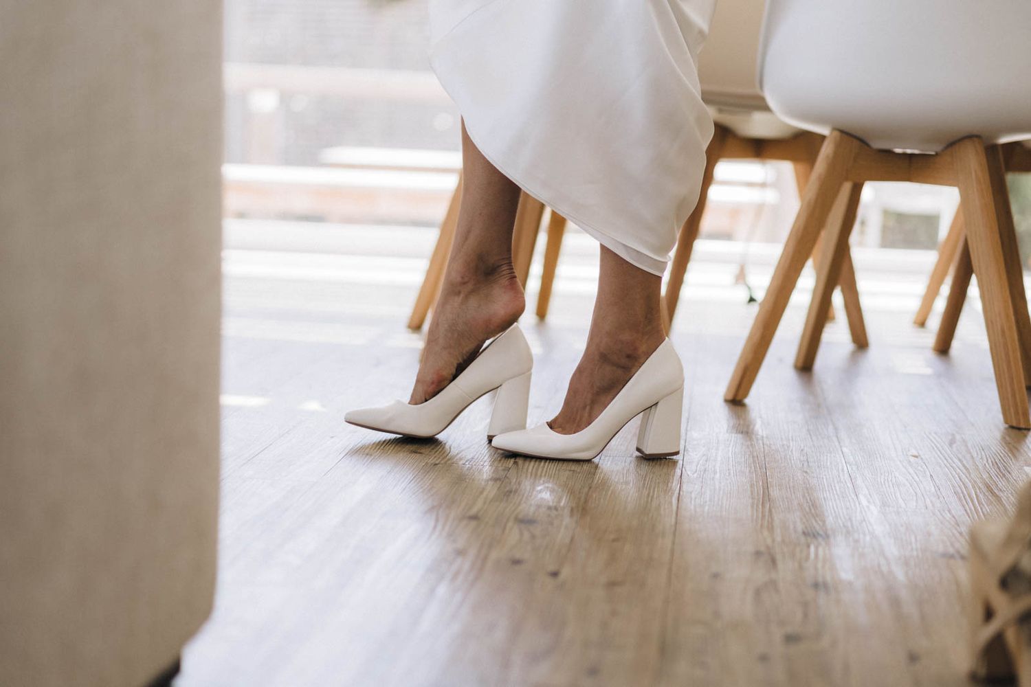 Elegant white block heel shoes peek out beneath a white dress on a hardwood floor beside wooden chairs.