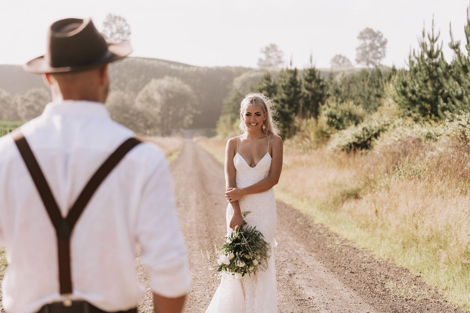 A bride in a white dress holds a bouquet on a sunlit dirt path as someone in suspenders looks on from the foreground.