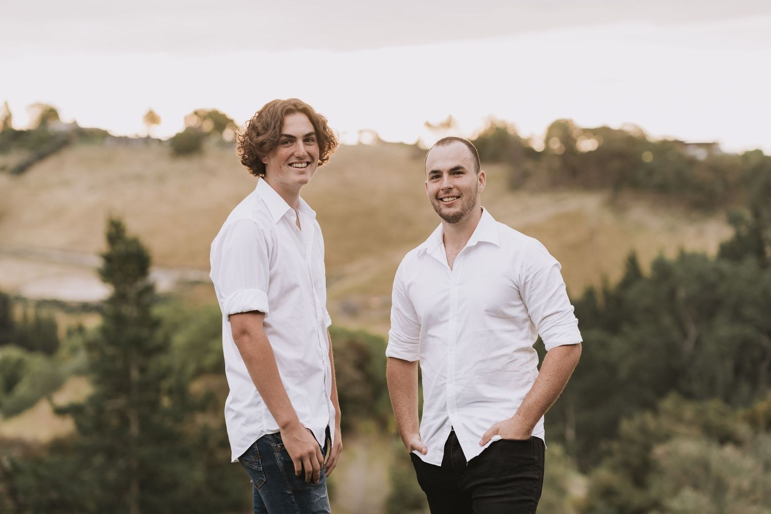 Two people in white button-down shirts stand together on a hilltop during golden hour with rolling hills in the background.