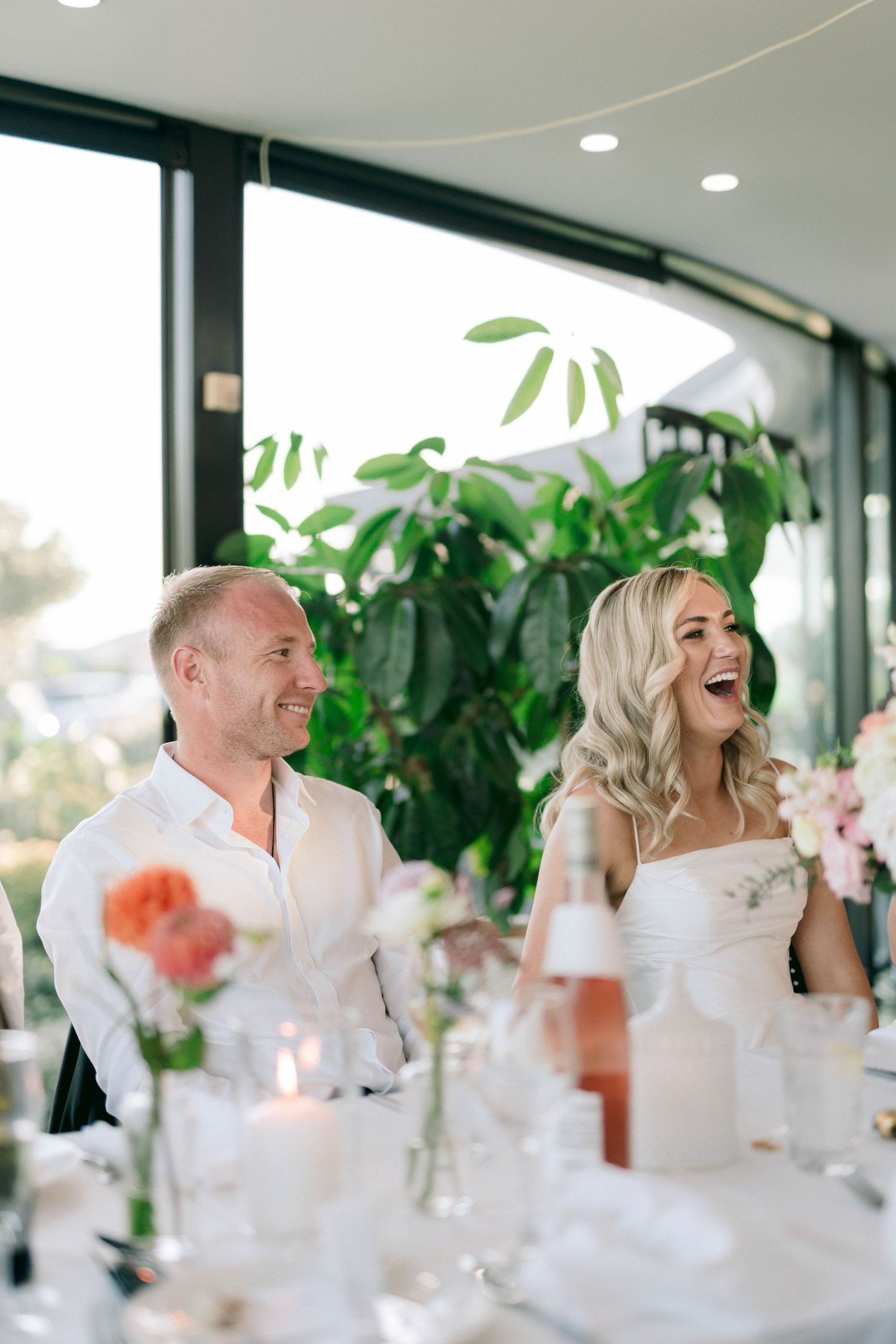 A wedding reception dinner scene with festive table settings, candlelight and indoor plants in the background.