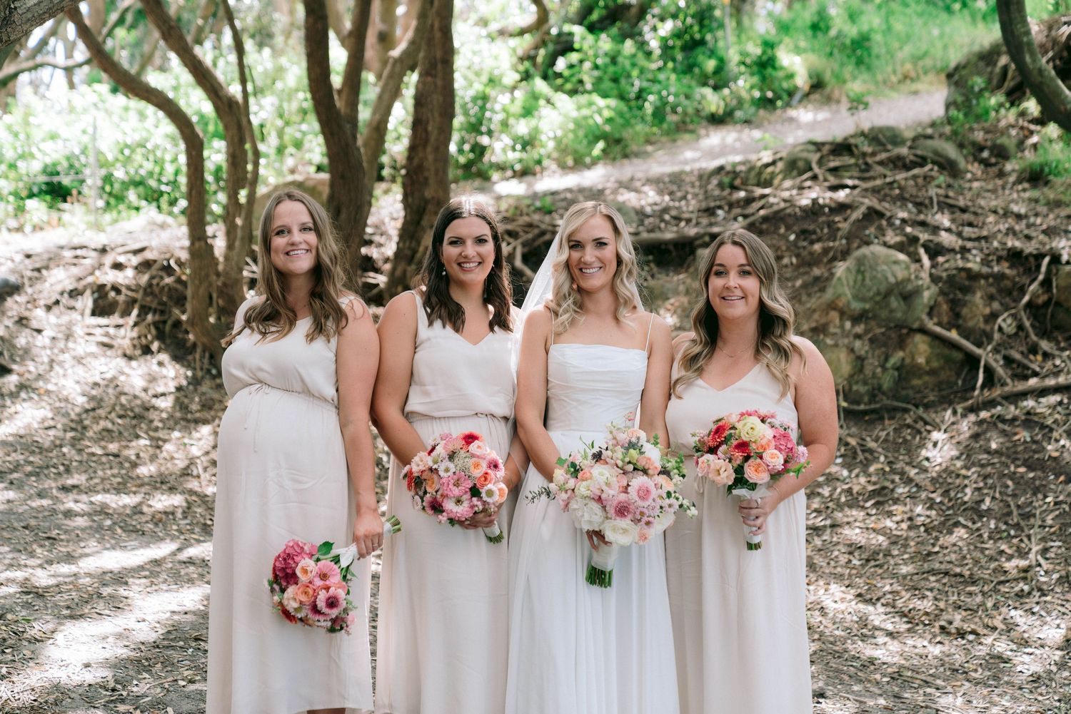 Four bridesmaids in light beige dresses stand together on a wooded path holding pink floral bouquets.
