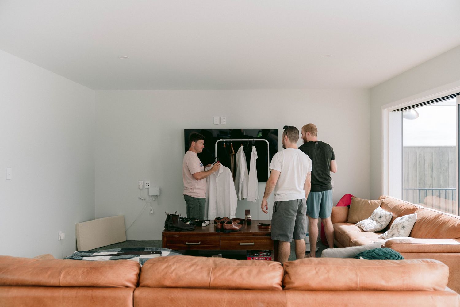 People gather around clothing rack in living room with tan leather sectional sofa and white walls.