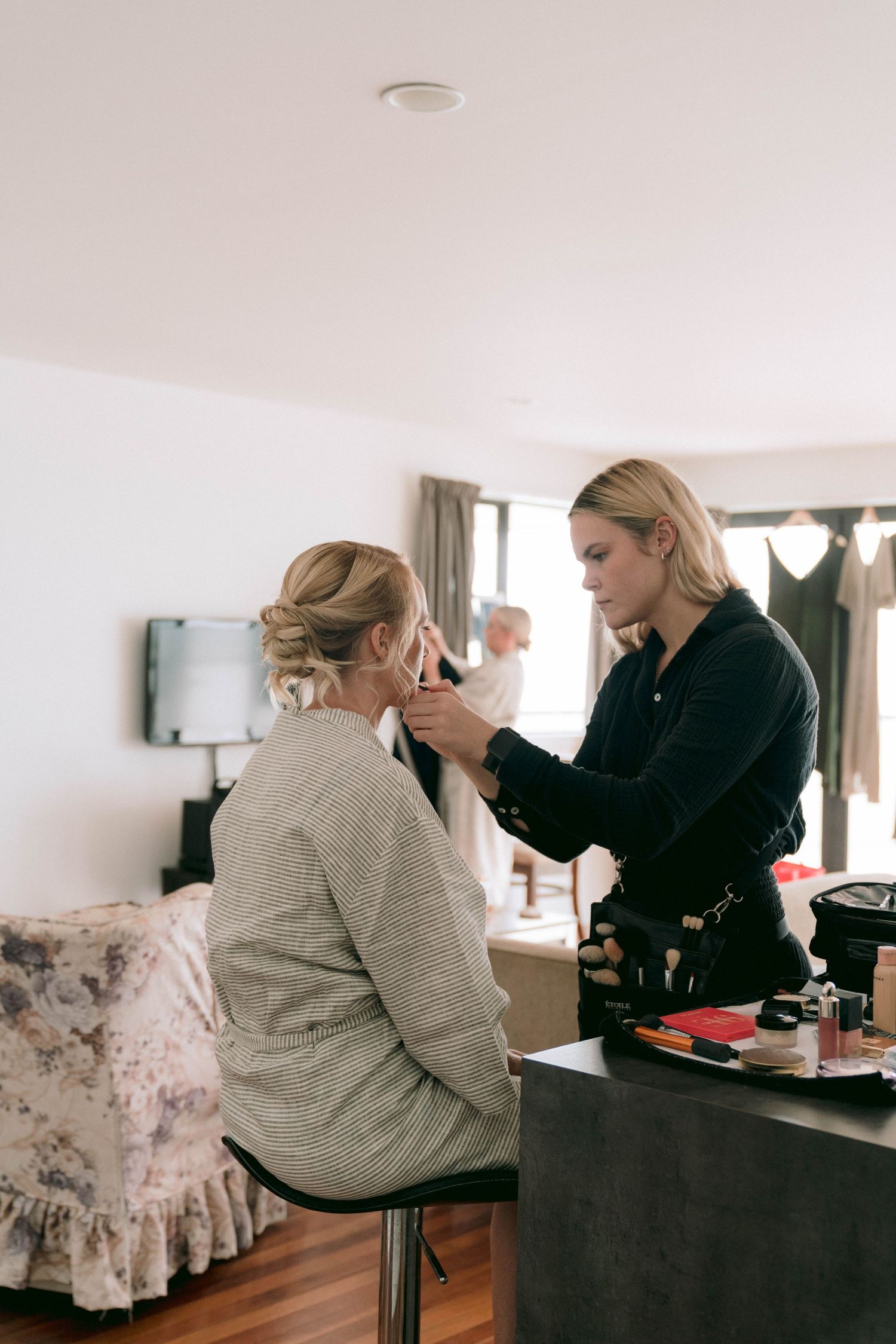 A makeup artist applies cosmetics to a client seated in a well-lit room with a floral chair.
