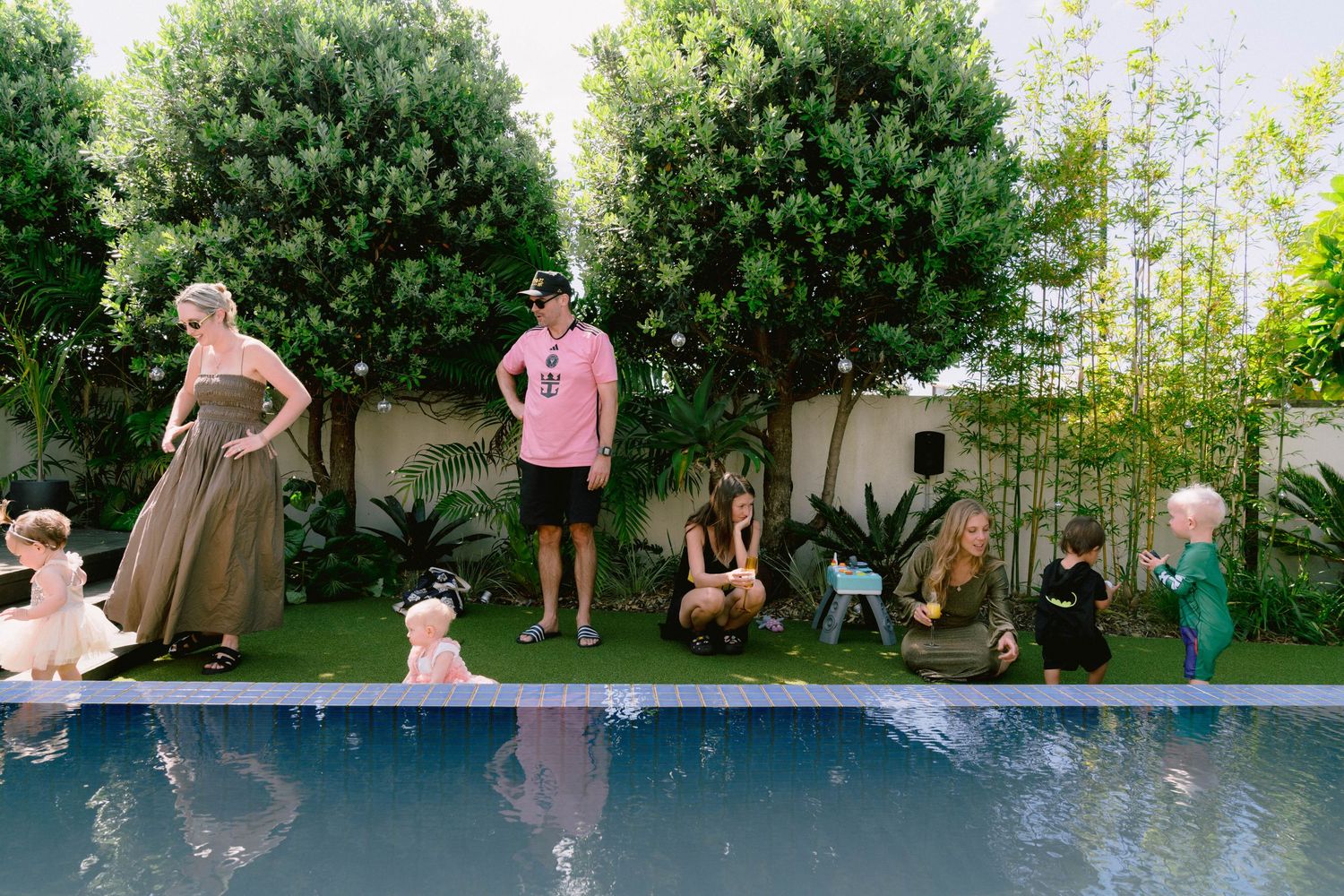 People gather around a backyard swimming pool surrounded by lush green trees on a sunny summer day.