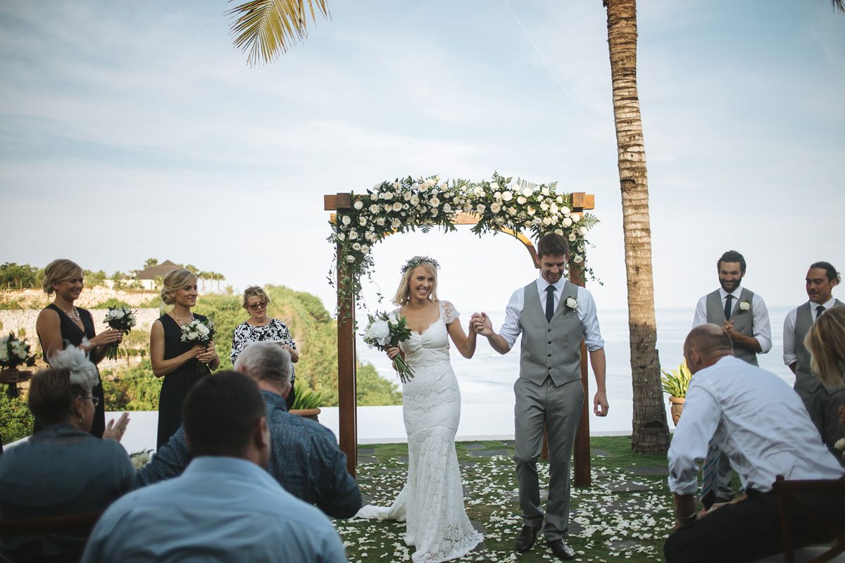 Wedding ceremony celebration under palm trees with guests and floral arch by the ocean.