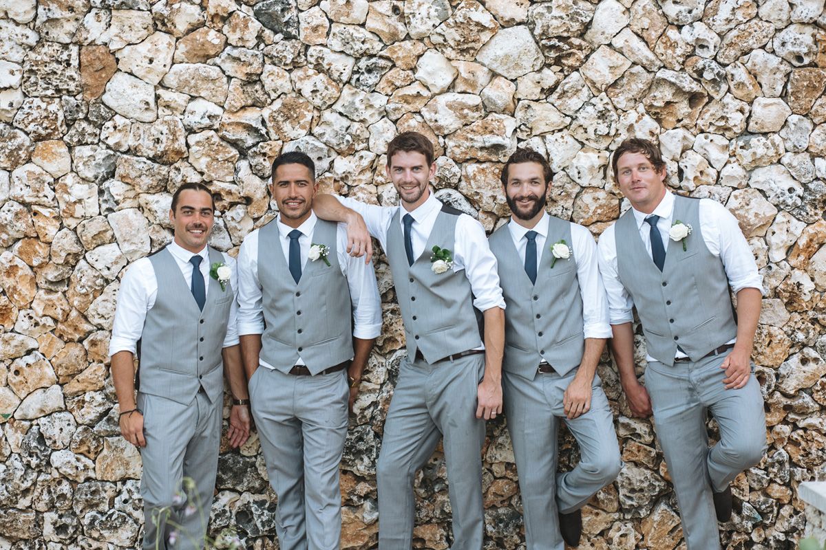 Groomsmen in light gray vests and suits pose against a stone wall background.