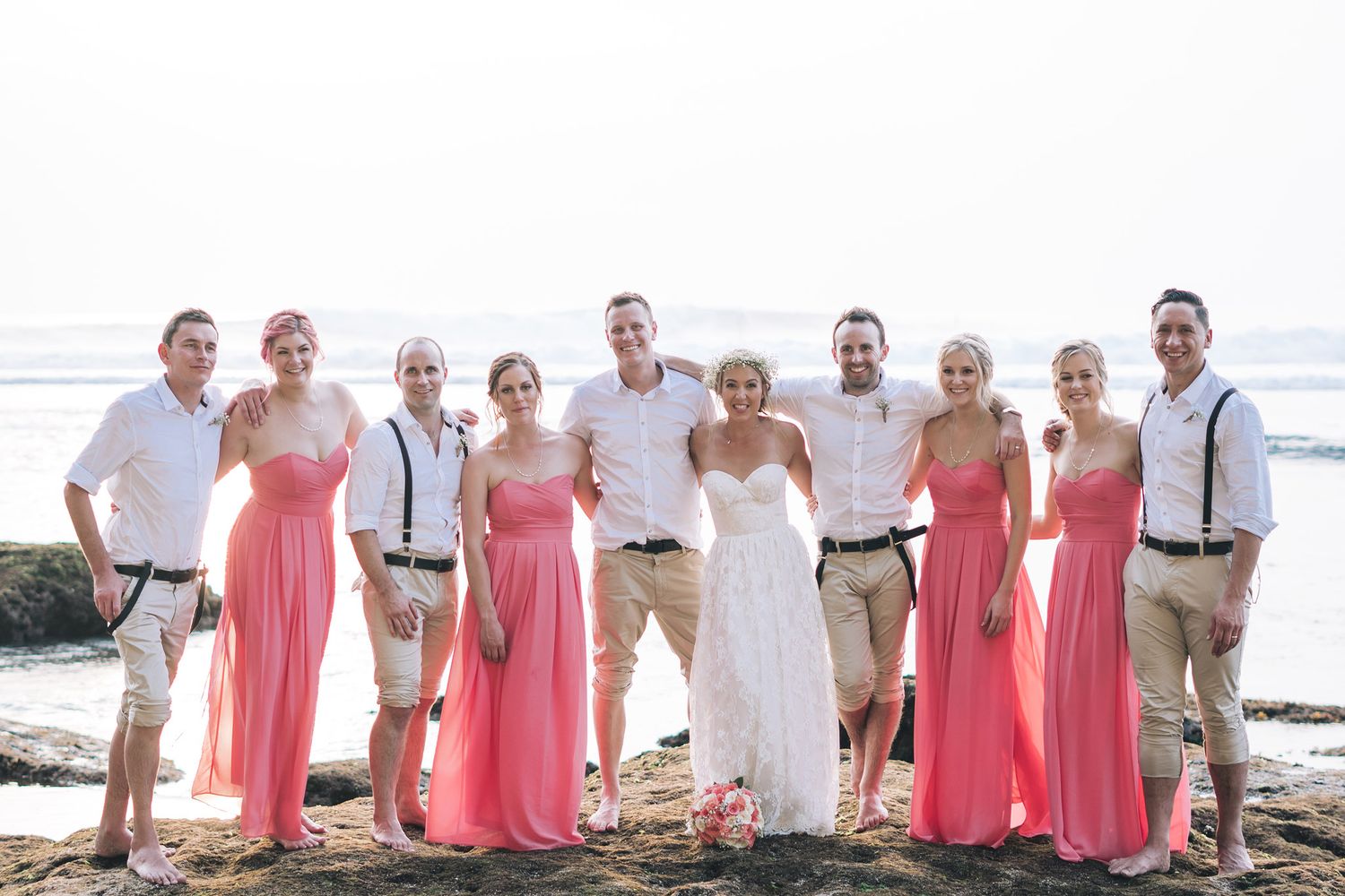 Wedding party in coral dresses and khaki pants posed together on rocky beach.
