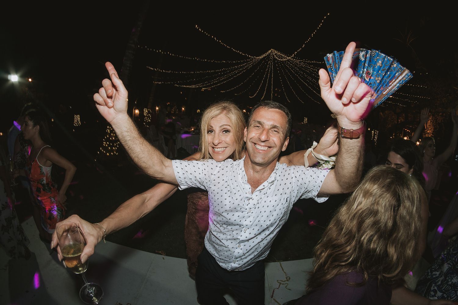 Dancing party guests celebrate on a dark dance floor with arms raised and lights glowing in background.