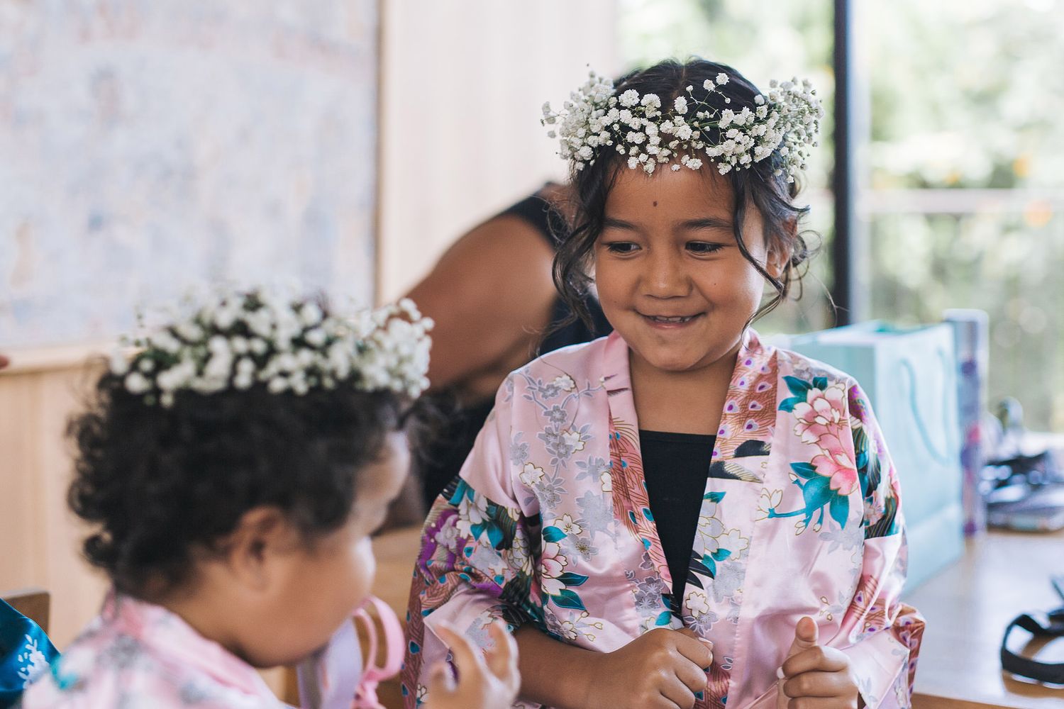 Children wearing floral robes and flower crowns share a joyful moment during preparation.