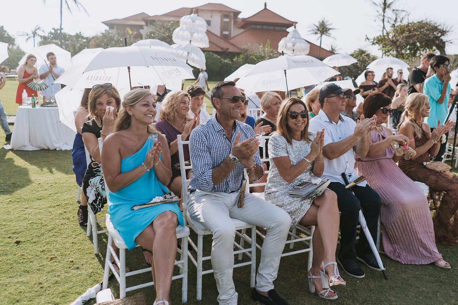 Wedding guests sit in white chairs holding parasols during an outdoor ceremony.