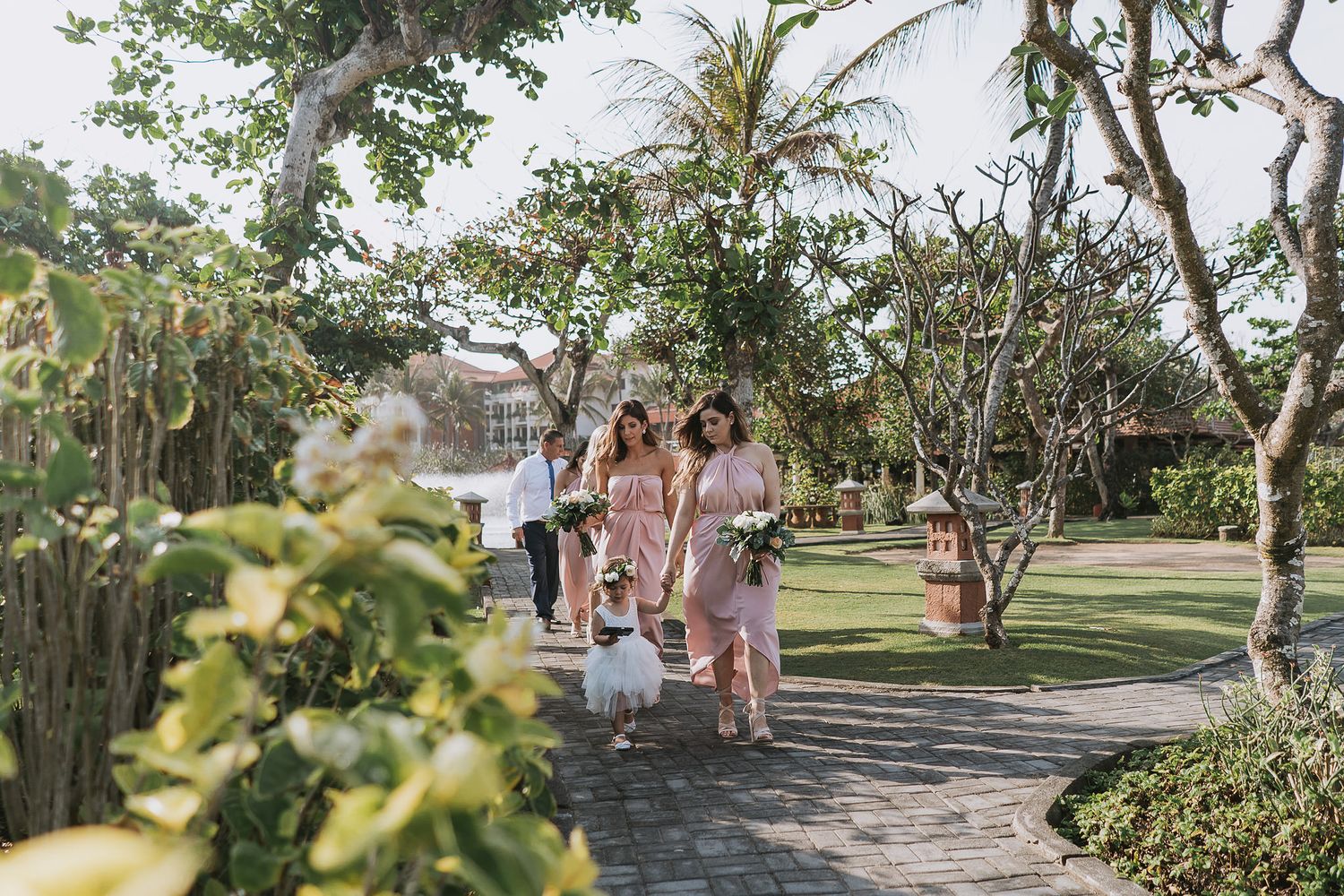 Wedding party walking along a palm tree-lined path in a garden setting.