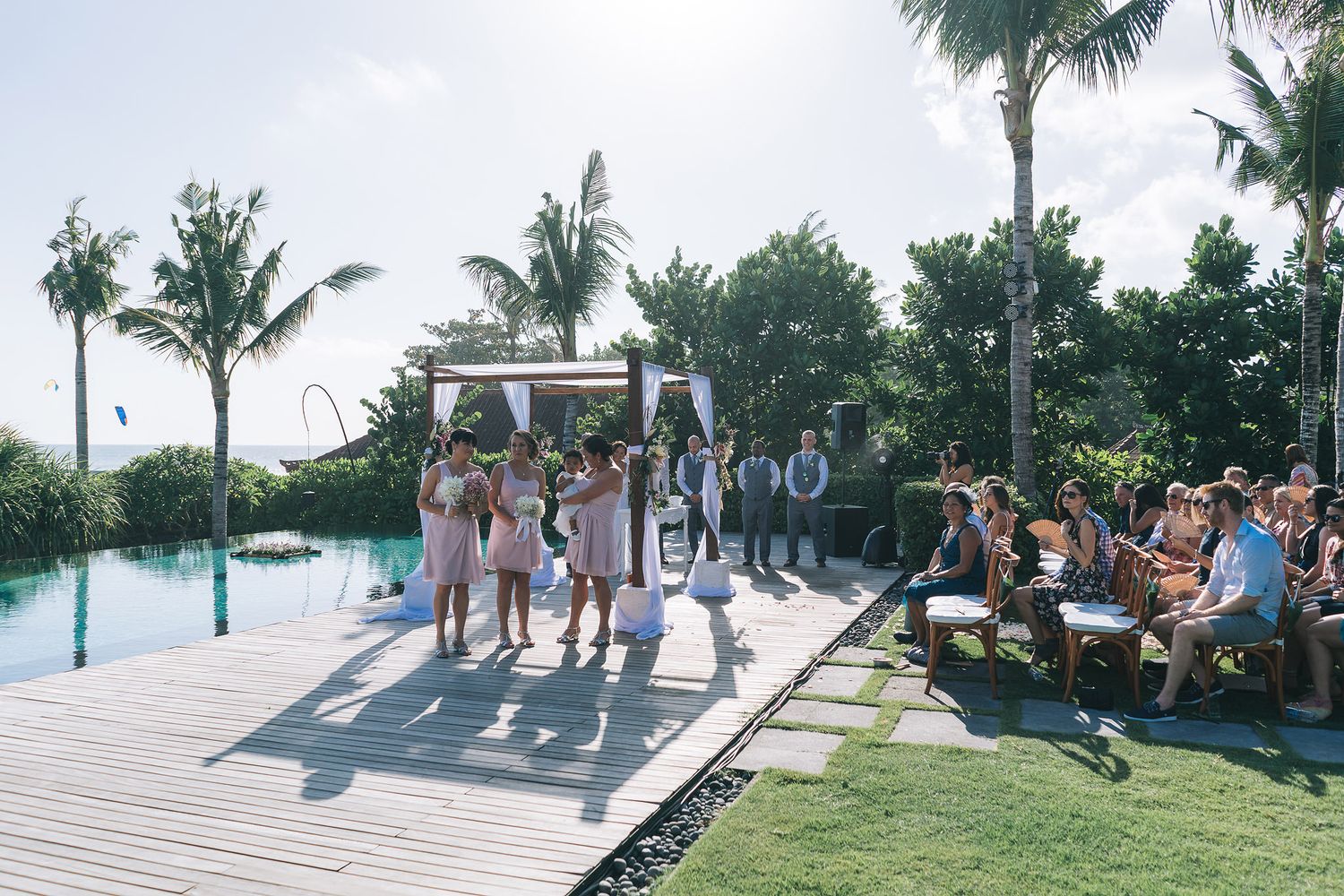 Palm trees line an outdoor wedding venue with white chairs set up on a lawn overlooking the ocean.