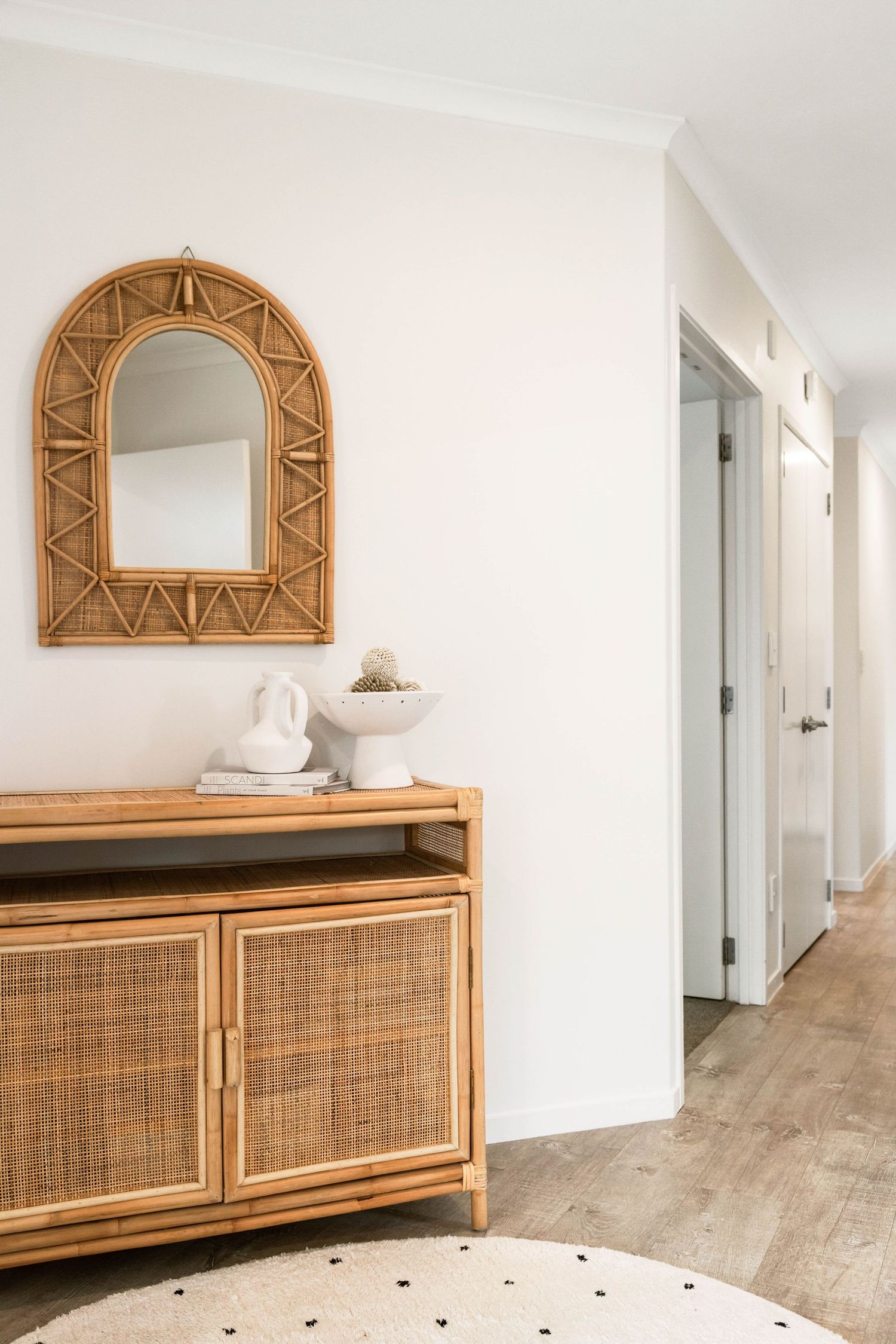 Modern rattan console table with sunburst mirror above in bright minimalist hallway with white walls and wood floors.