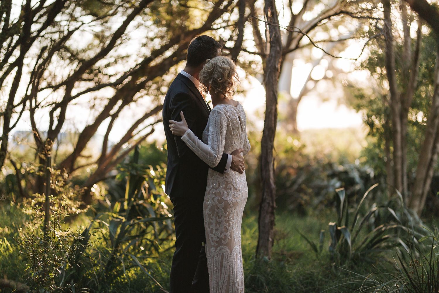 A couple embraces during golden hour in a lush garden setting with sunlight filtering through the trees.