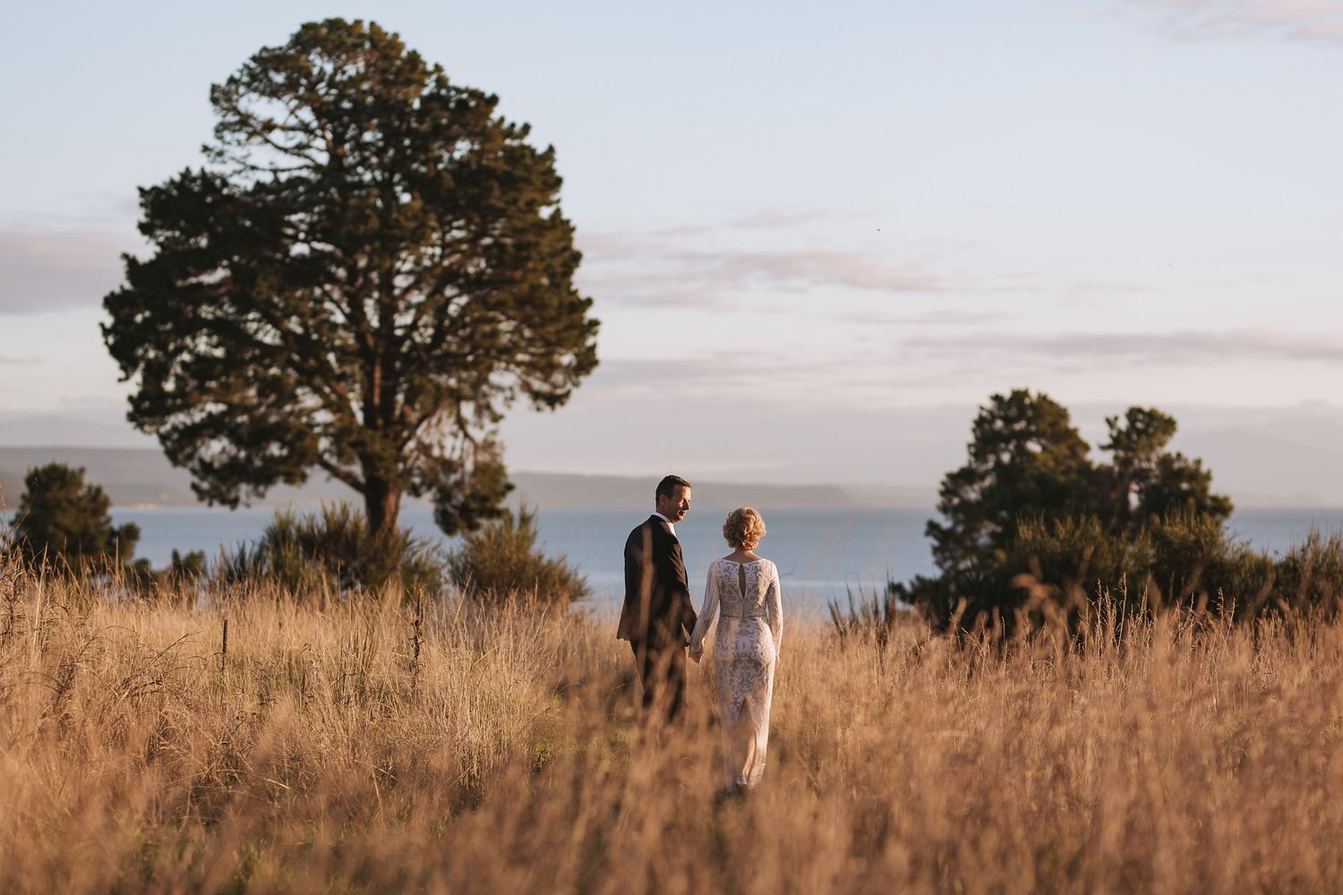 Two figures walk together through tall golden grass at sunset with a large tree silhouetted against the sky.