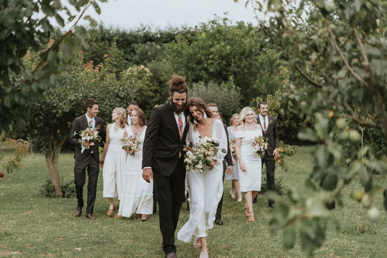 A wedding party walks through a green garden path during an outdoor countryside celebration.