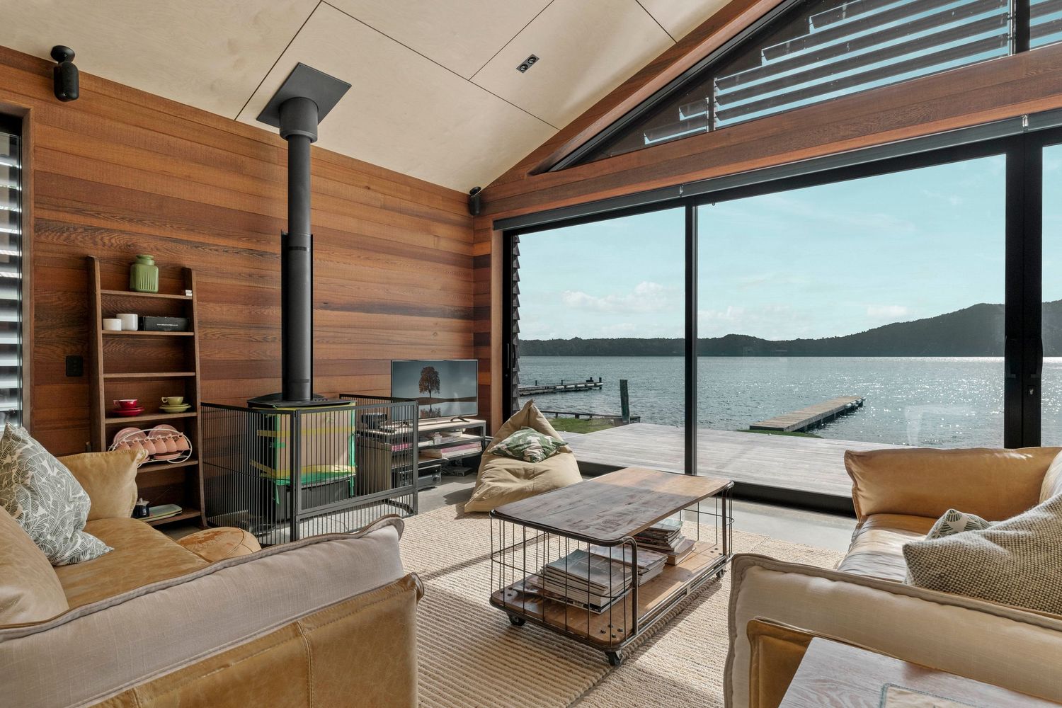 Modern coastal living room with panoramic water views, wood paneling, and a wood-burning stove beside comfortable seating.