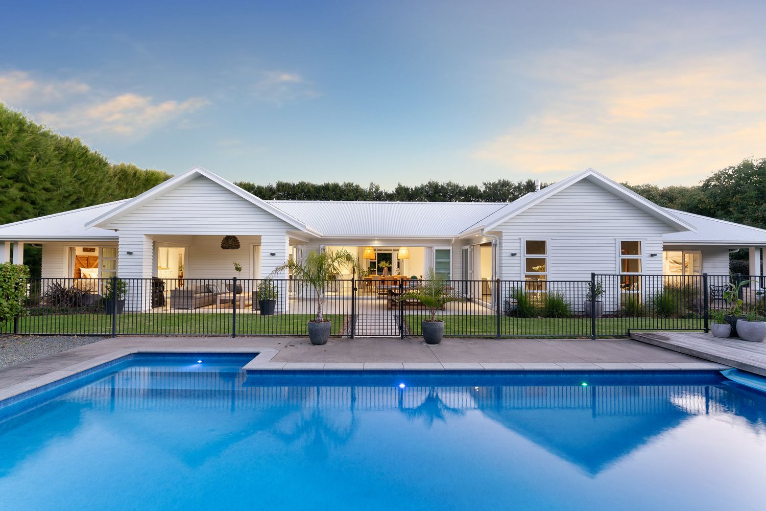 Luxurious white ranch-style home with swimming pool, deck and black metal fencing in tropical setting.