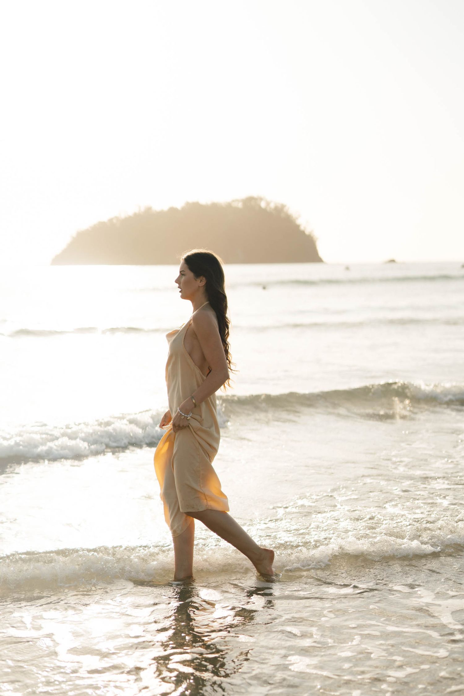 Person in light dress splashes through ocean waves during sunset beach session.
