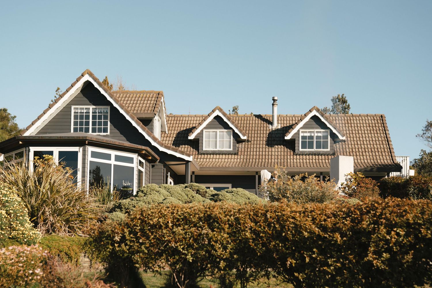 Large craftsman style home with grey siding and multiple dormers sits atop a hill surrounded by autumn foliage.