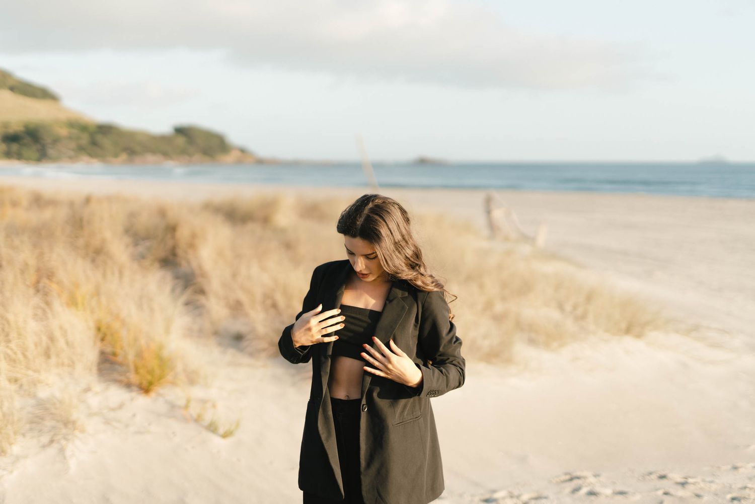 Figure dressed in black stands on a sandy beach with golden grass and ocean waves in the background during sunset.