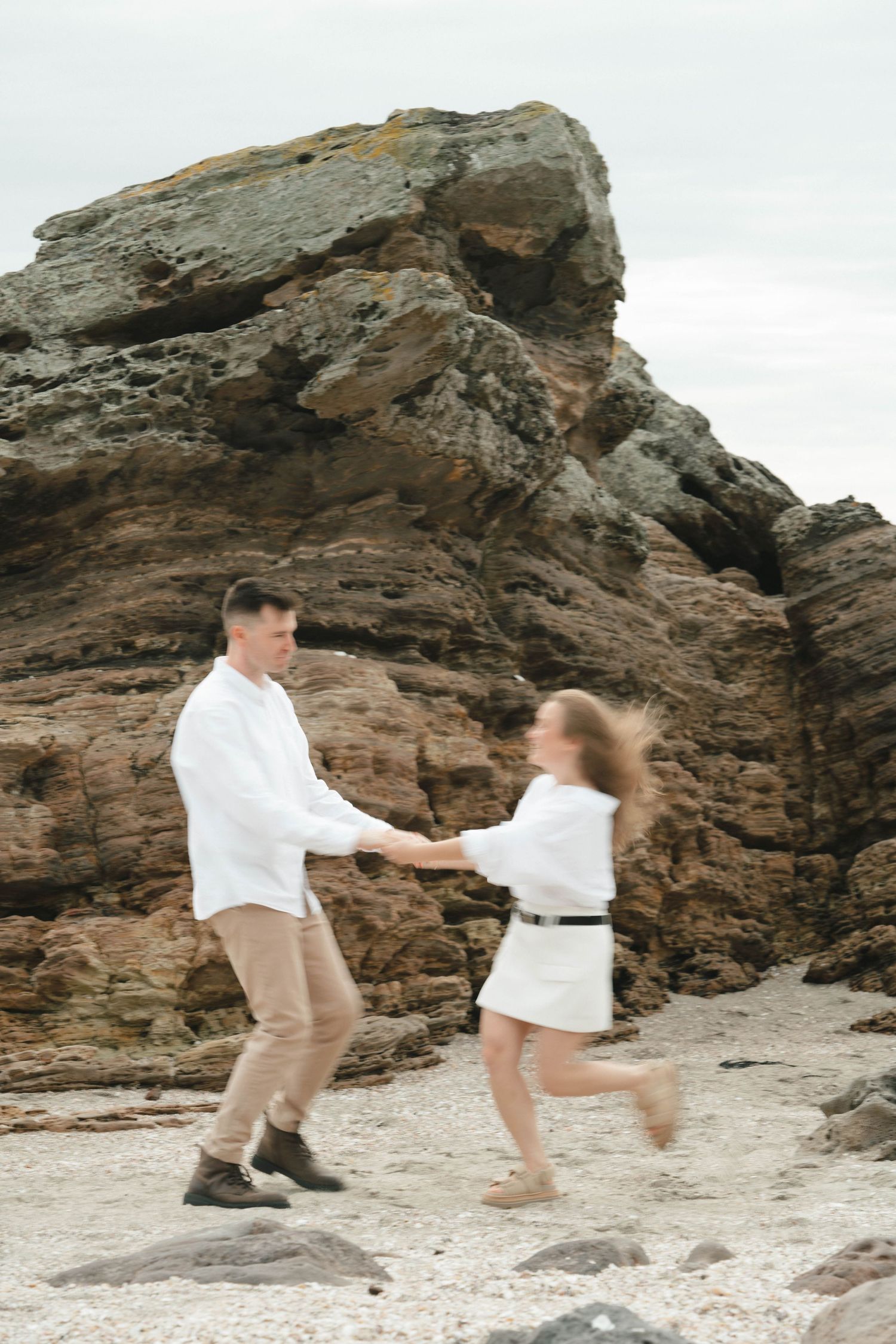 A couple dances playfully on a beach with dramatic cliffs behind them.