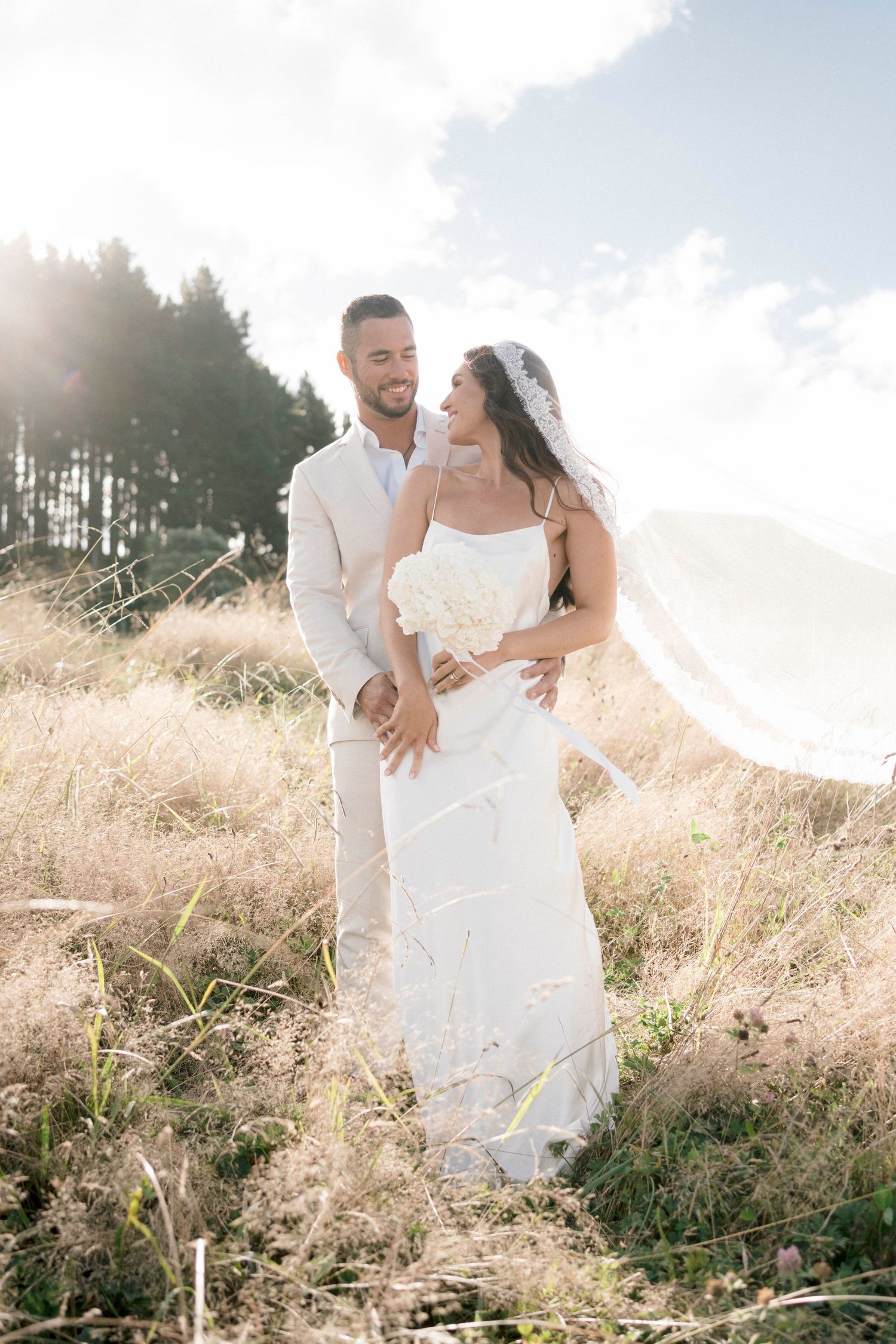 Happy couple in white wedding attire share intimate moment in sunlit field with pine trees.