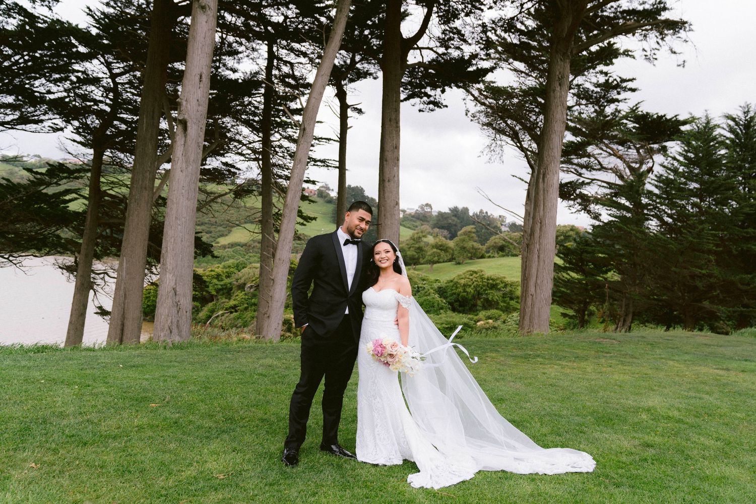 A wedding couple stands together on a grassy lawn surrounded by tall evergreen trees near the coast.