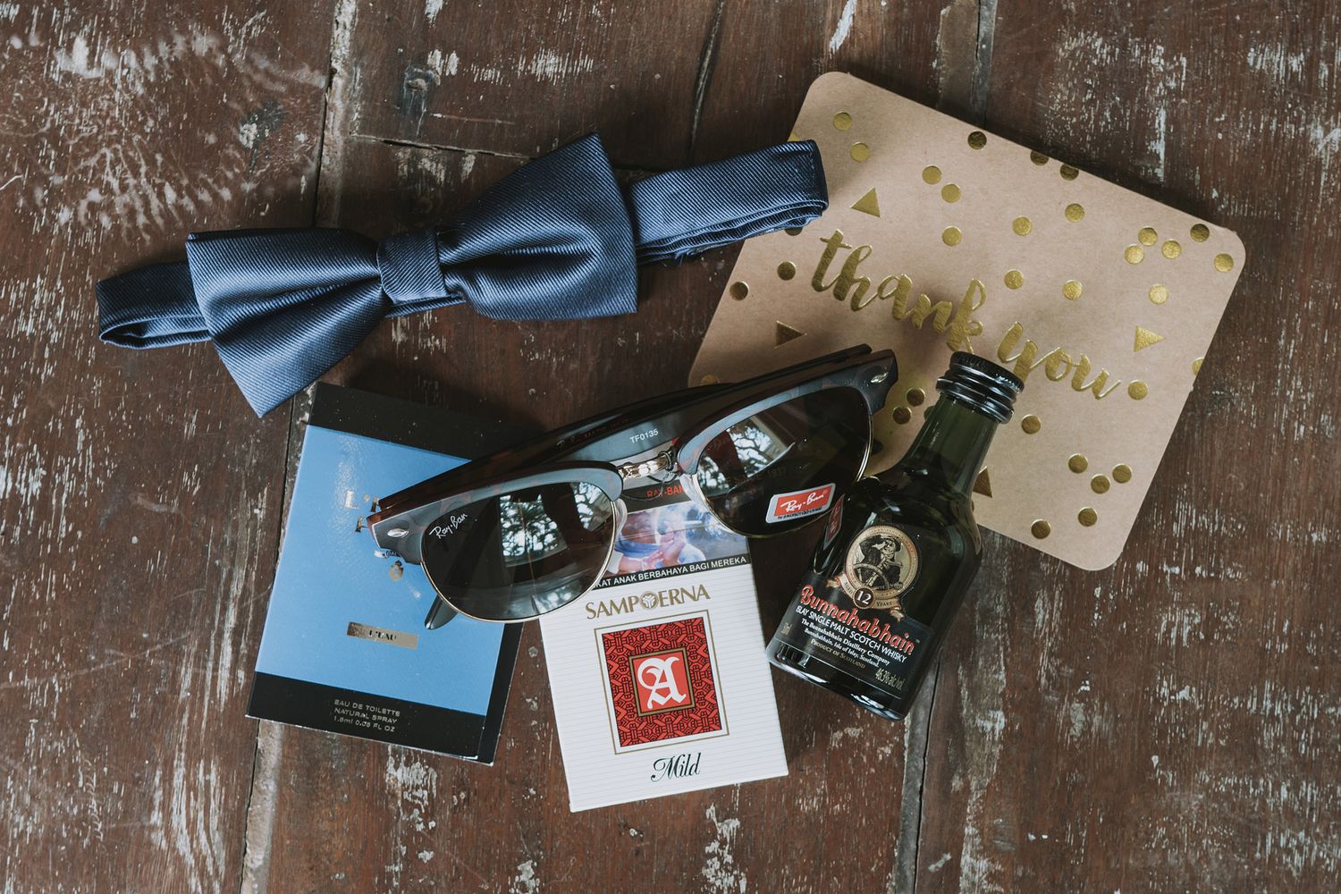 Wedding accessories including a blue bowtie, cards and a glass bottle arranged on rustic wooden surface.