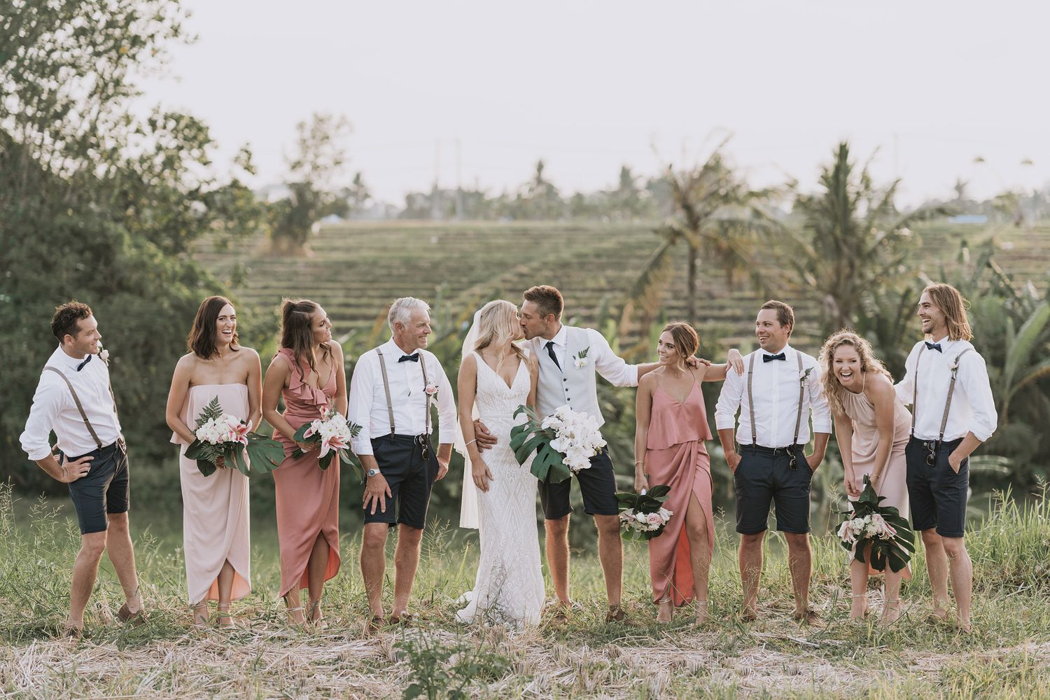 A wedding party lined up outdoors in pink dresses and black suits against a natural landscape.