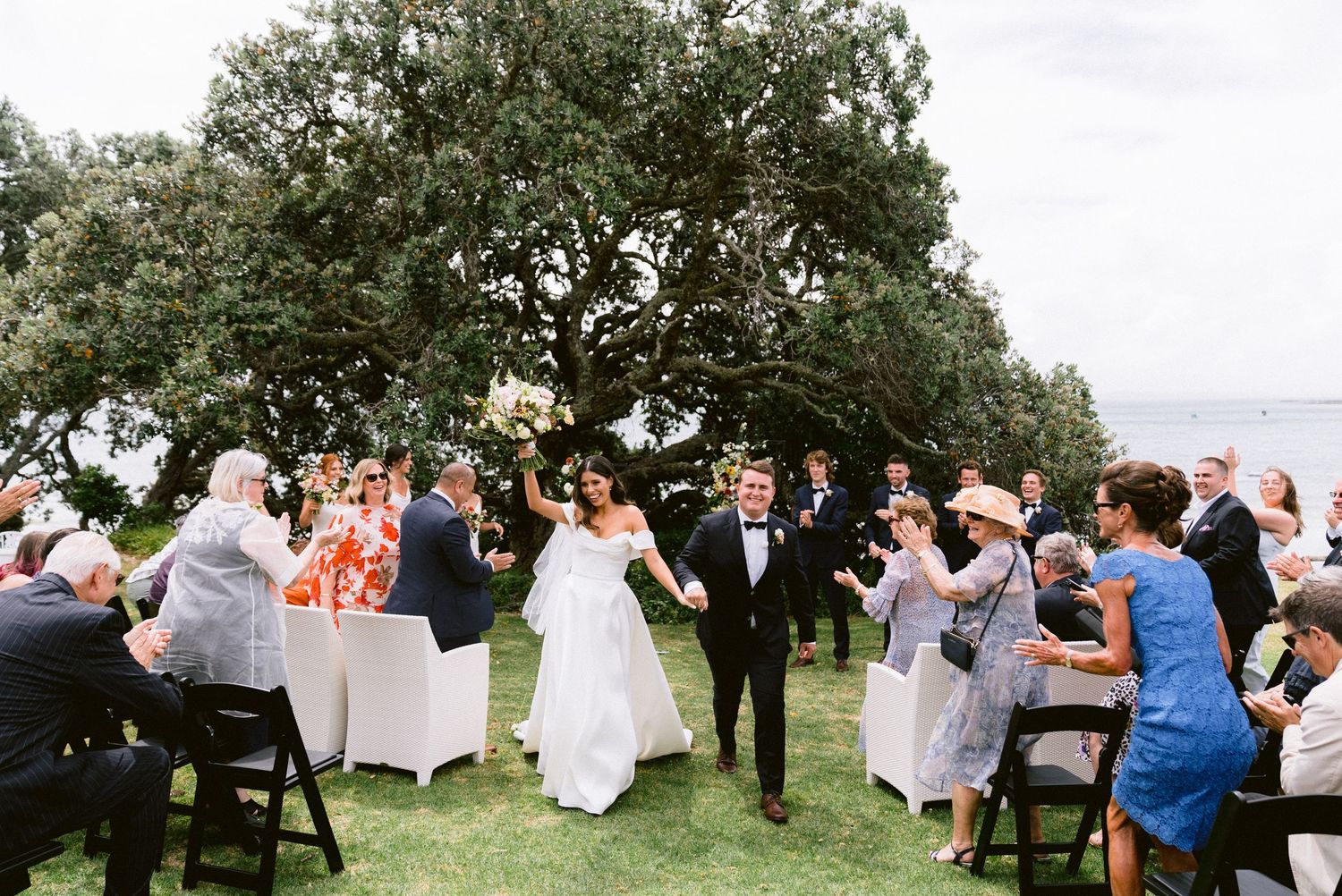 Wedding guests seated on a grassy lawn during an outdoor ceremony with trees in the background.