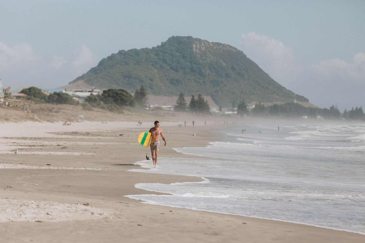 A lone surfer walks along a misty beach with a mountain peak in the background.