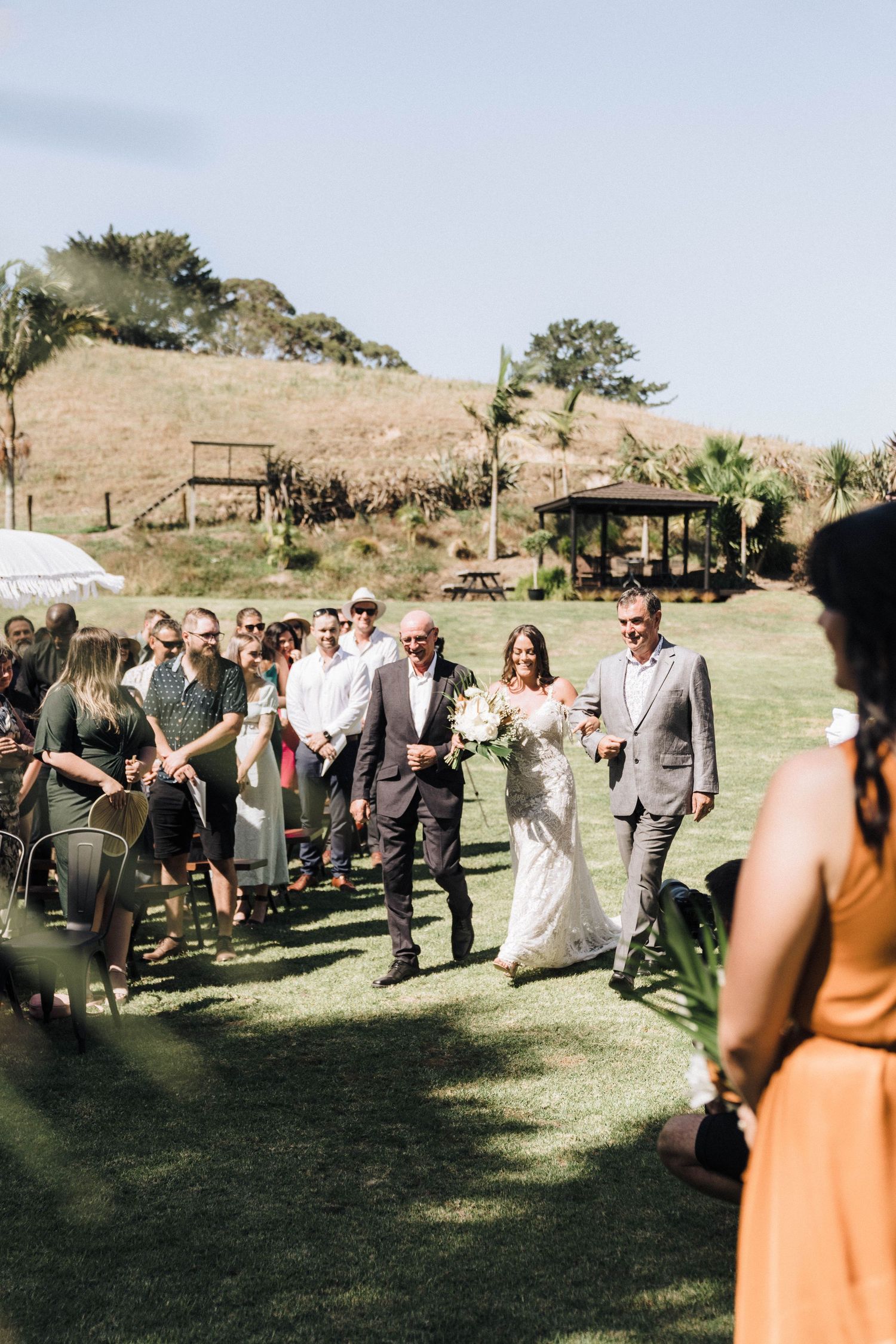 Wedding ceremony guests gather on a grassy lawn with hills and gazebo in the background on a sunny day.