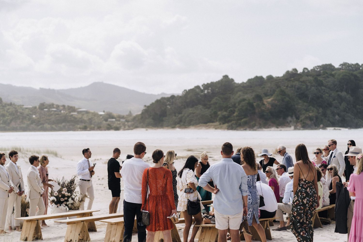 A group of people gather on a wooden deck overlooking a lake surrounded by mountains on a cloudy day.
