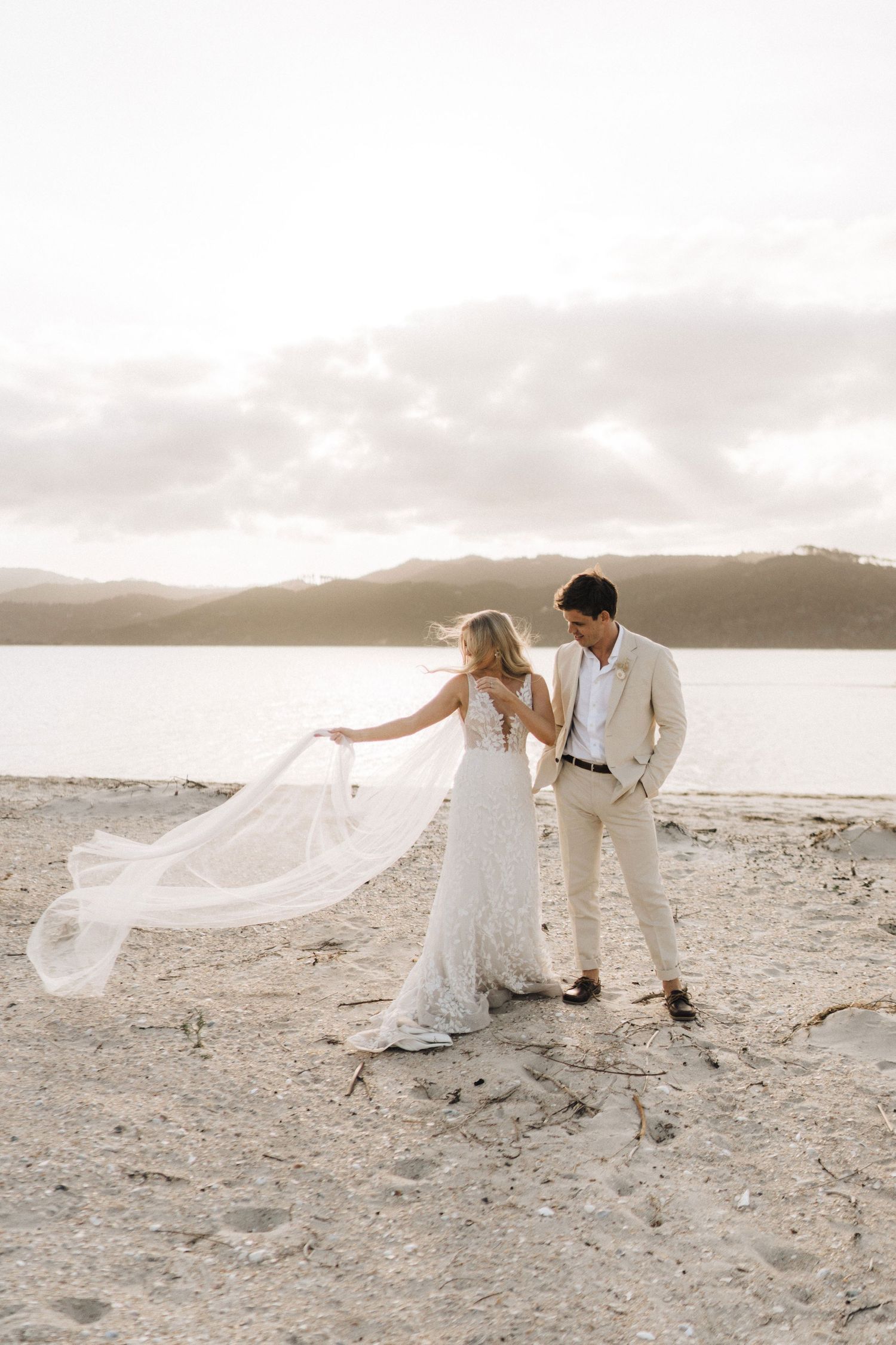 Romantic beach wedding couple in white standing by the ocean as bride's veil flows dramatically in the wind across sandy shore.