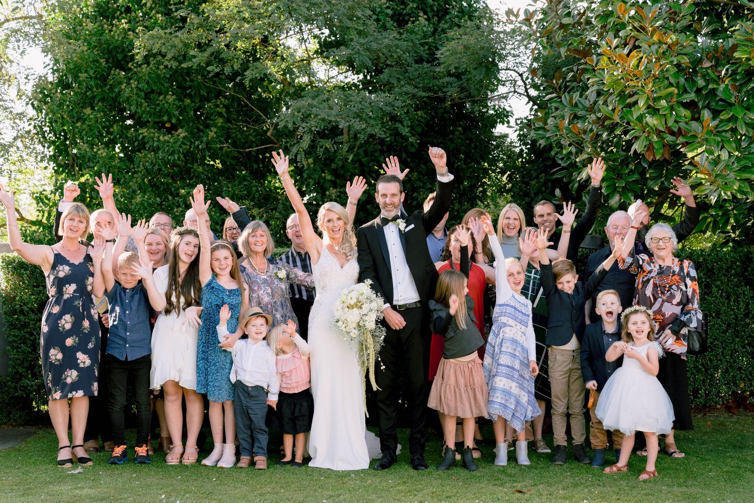 Large group of wedding guests gathered outdoors in a garden setting for celebratory photos.