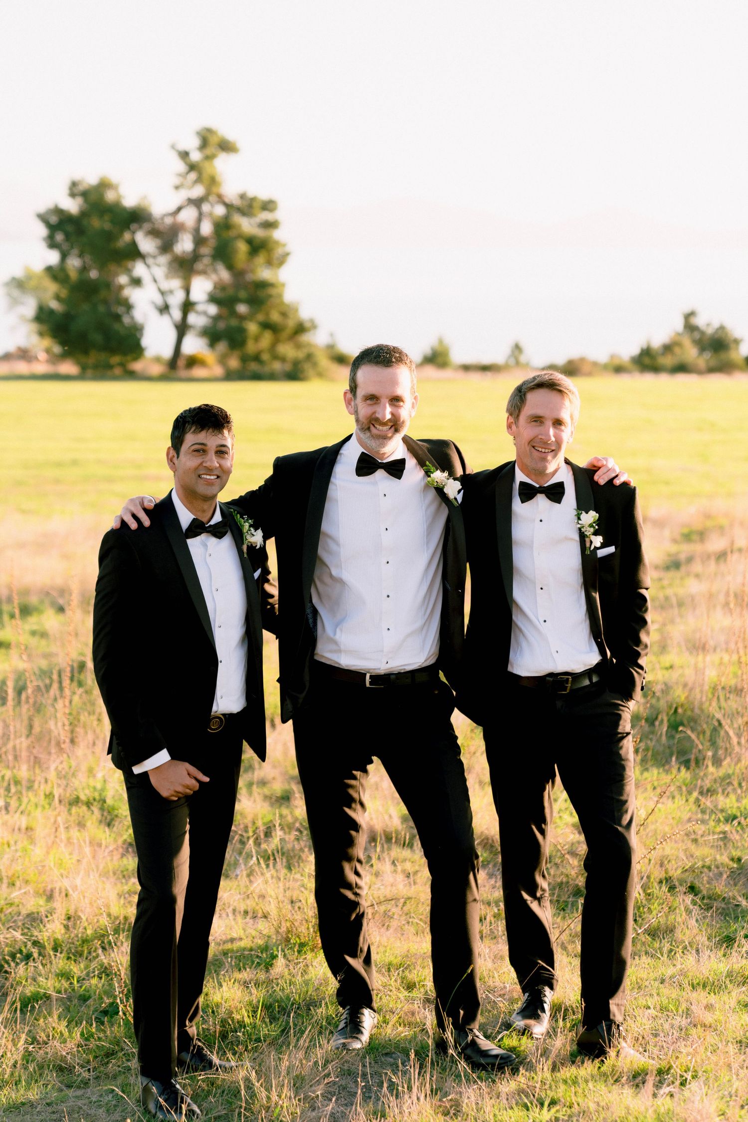 Three groomsmen in black tuxedos standing together in golden field at sunset.