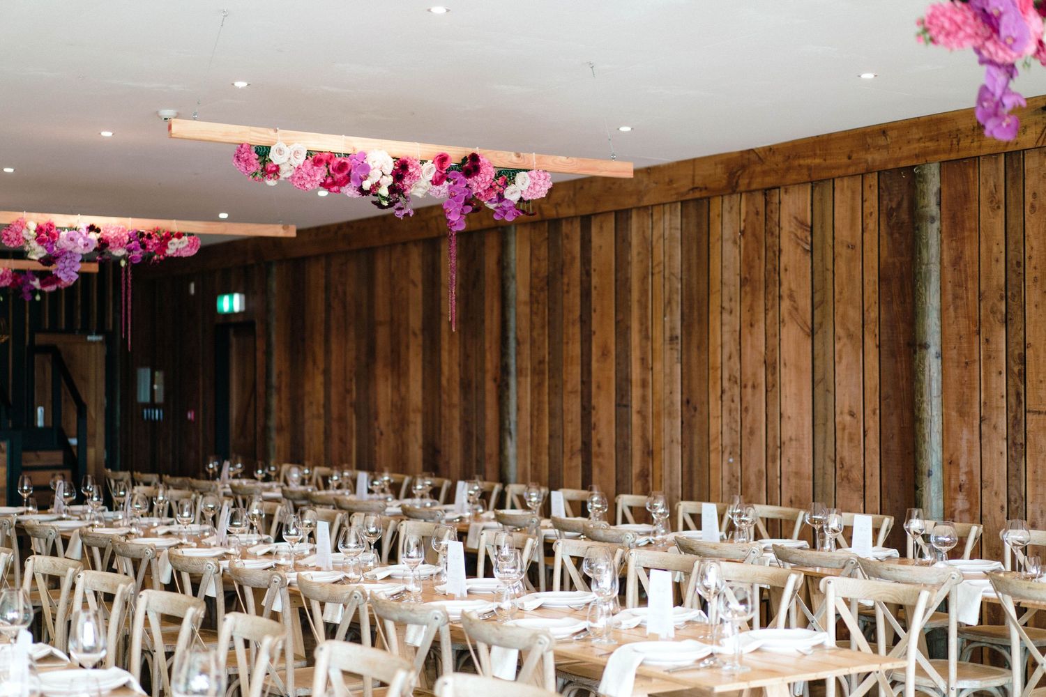 Reception venue with wooden walls, white cross-back chairs, and pink floral ceiling installations.